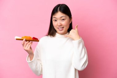 Elinde saşimi olan genç bir Çinli kadın pembe arka planda izole edilmiş bir şekilde telefon jesti yapıyor. Bana geri dönüş işareti ver.