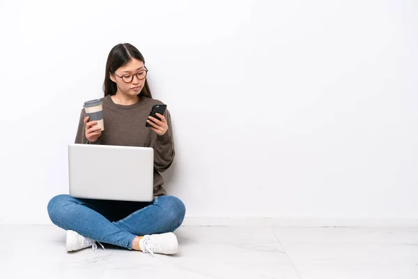 一个拿着笔记本电脑的年轻女人坐在地板上 手里拿着咖啡 一边拿着手机 — 图库照片