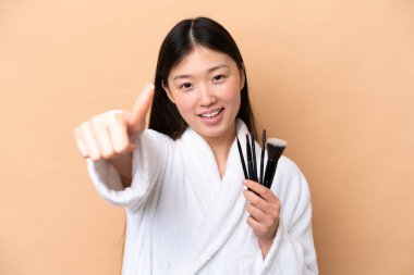 Genç Çinli kadın bej arka planda izole edilmiş elinde makyaj fırçası ve başparmağıyla