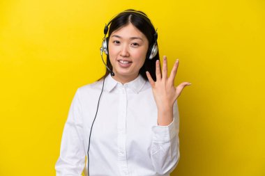 Telefon pazarlamacısı Çinli kadın sarı arka planda izole edilmiş kulaklıkla çalışıyor. Beş parmaklı sayım yapıyor.