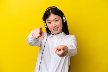 Sarı arka planda izole edilmiş kulaklıkla çalışan pazarlamacı Çinli kadın sana gülümserken parmak atıyor.