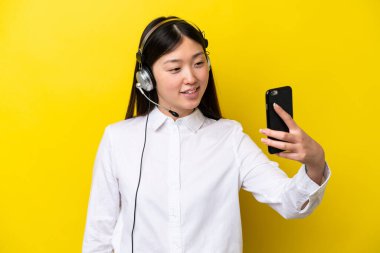 Tele pazarlamacı Çinli kadın sarı arka planda izole edilmiş bir kulaklıkla selfie çekiyor.