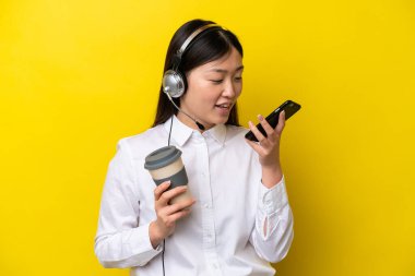Tele pazarlamacı Çinli bir kadın sarı arka planda izole edilmiş bir kulaklıkla çalışıyor. Elinde kahve ve cep telefonu var.
