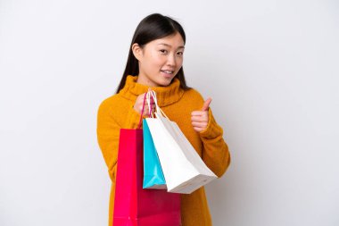 Beyaz arka planda izole edilmiş genç bir Çinli kadın elinde alışveriş çantaları ve başparmağıyla