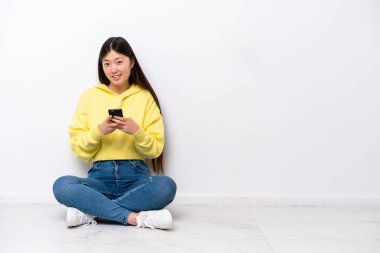 Genç Çinli bir kadın beyaz duvarda izole bir şekilde yerde oturuyor ve cep telefonuyla bir mesaj gönderiyor.