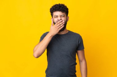 Genç Faslı bir adam sarı arka planda izole edilmiş esniyor ve ağzı açık bir şekilde elle kapanıyor.