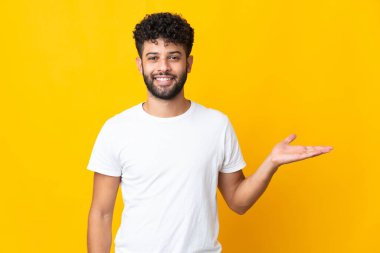 Faslı genç bir adam sarı arka planda izole edilmiş. Elinde bir ilan vermek için avucunda hayali bir alan tutuyor.