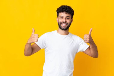 Faslı genç adam sarı arka planda izole edilmiş bir şekilde baş parmak hareketi yapıyor.