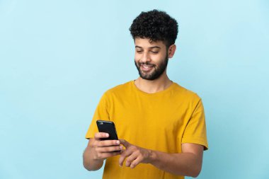 Genç Faslı bir adam mavi arka planda izole edilmiş. Cep telefonuyla mesaj ya da e-posta gönderiyor.