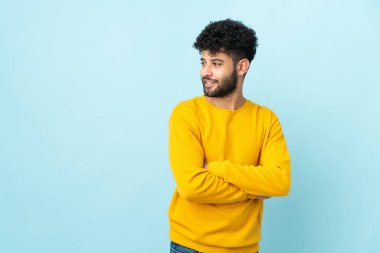 Faslı genç bir adam mavi arka planda izole edilmiş, yana bakıyor ve gülümsüyor.