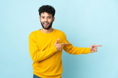 Faslı genç bir adam mavi arka planda izole edilmiş. Şaşırmış ve işaret ediyor.