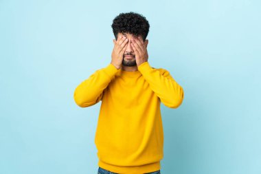 Faslı genç bir adam mavi arka planda izole edilmiş. Gözlerini elleriyle kapatıyor.