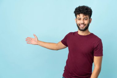 Faslı genç bir adam mavi arka planda izole edilmiş. Davet etmek için ellerini yana uzatıyor.