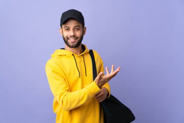 Spor çantalı genç Faslı adam mor arka planda izole edilmiş. Davet etmek için ellerini yana uzatıyor.
