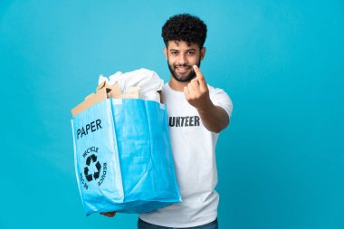 Genç Faslı bir adam, izole edilmiş arkaplanı geri dönüştürmek için içi kağıt dolu bir geri dönüşüm çantası tutuyor.