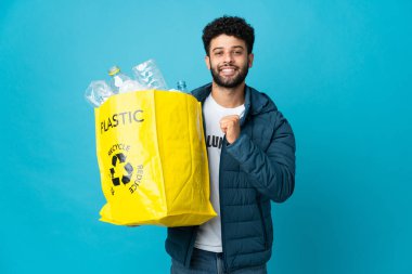 Genç Faslı bir adam elinde plastik şişelerle dolu bir çanta ile izole edilmiş arka planda gururlu ve kendini beğenmiş bir şekilde geri dönüşüm yapıyor.