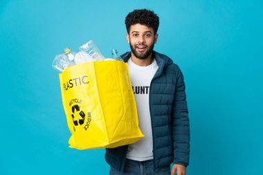 Genç Faslı bir adam, izole edilmiş arkaplanı süpriz bir yüz ifadesiyle geri dönüştürmek için plastik şişelerle dolu bir çanta tutuyor.