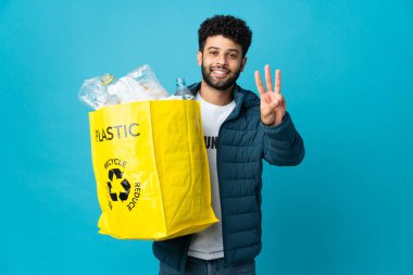 Genç Faslı bir adam izole edilmiş arka planda geri dönüşüm yapmak için plastik şişelerle dolu bir çanta tutuyor mutlu ve parmaklarıyla üçe kadar sayıyor.