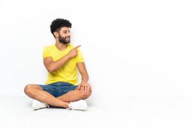 Genç Faslı yakışıklı bir adam izole bir zeminde oturuyor ve bir ürünü sunmak için yan tarafı işaret ediyor.