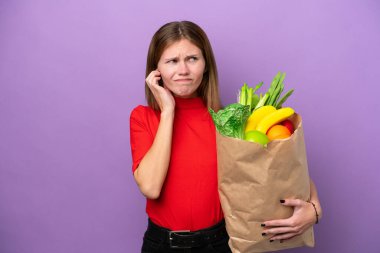 Mor arka planda izole edilmiş alışveriş torbası tutan genç İngiliz kadın hayal kırıklığına uğramış ve kulaklarını kapatmış.