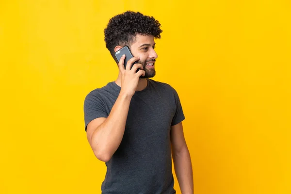 Young Moroccan man isolated on yellow background keeping a conversation with the mobile phone with someone