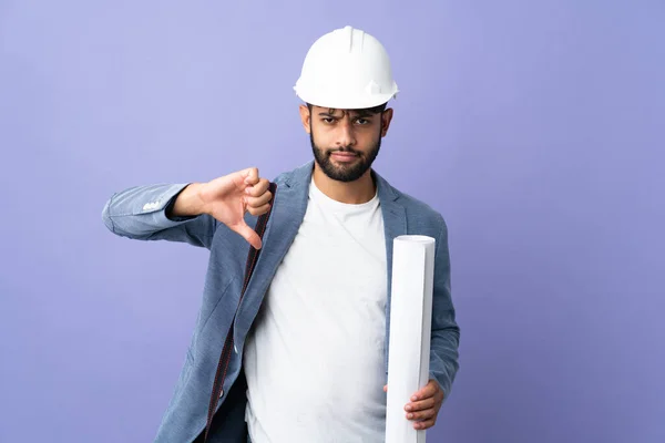 Young architect Moroccan man with helmet and holding blueprints over isolated background showing thumb down with negative expression