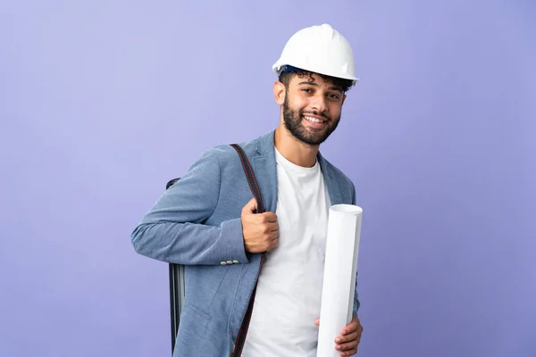 Young architect Moroccan man with helmet and holding blueprints over isolated background laughing