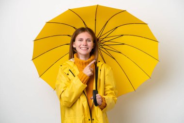 Yağmura dayanıklı ceketli ve şemsiyeli genç bir İngiliz kadın beyaz arka planda izole edilmiş bir ürün sunmak için yan tarafı işaret ediyor.