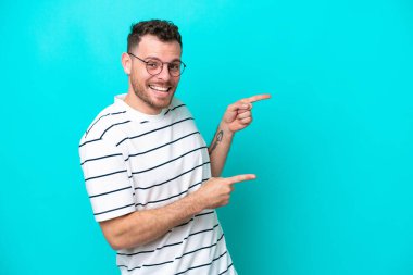 Mavi arka planda izole edilmiş genç Brezilyalı adam şaşırmış ve işaret ediyor.