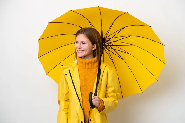 若いです英国人女性でレインプルーフコートと傘は白い背景に隔離されました側を見て笑顔 — ストック写真