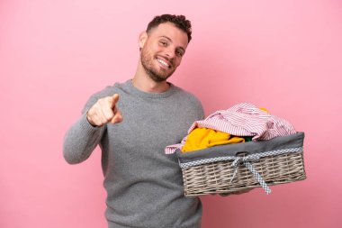 Genç Brezilyalı bir adam pembe arka planda izole edilmiş bir sepet tutuyor mutlu bir ifadeyle ön tarafı işaret ediyor.