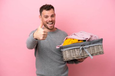 Genç Brezilyalı bir adam pembe arka planda izole edilmiş bir çamaşır sepeti tutuyor. Başparmakları havada çünkü iyi bir şey oldu.