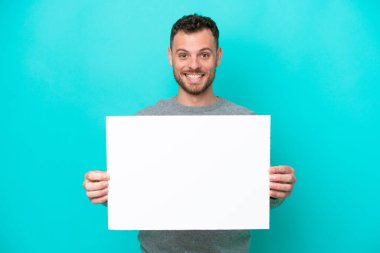 Elinde boş bir levha tutan genç Brezilyalı adam mavi arka planda izole edilmiş mutlu bir ifadeyle boş bir pankart tutuyor.