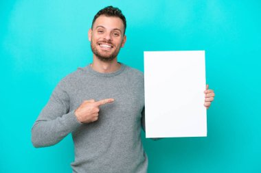 Genç Brezilyalı bir adam elinde boş bir levha tutuyor mavi arka planda izole edilmiş mutlu bir ifadeyle boş bir levha tutuyor ve onu işaret ediyor.