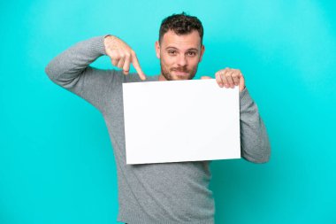 Genç Brezilyalı bir adam elinde boş bir levha tutuyor mavi arka planda izole edilmiş mutlu bir ifadeyle boş bir levha tutuyor ve onu işaret ediyor.