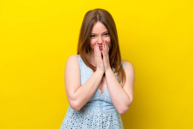 Sarı arka planda izole edilmiş genç beyaz kadın mutlu ve gülümseyen elleriyle ağzını kapatıyor.