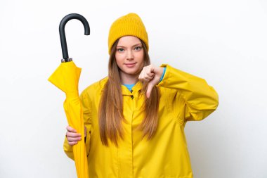 Yağmura dayanıklı ceketi ve şemsiyesi olan beyaz arka planda izole genç beyaz kadın, olumsuz bir ifadeyle baş parmağını aşağı gösteriyor.