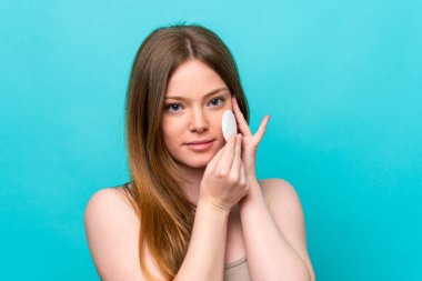 Yüzündeki makyajı silmek için mavi arka planda pamuklu pedi olan beyaz bir kadın.