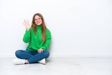 Beyaz arka planda yerde oturan beyaz tenli genç kadın parmaklarıyla beşe kadar sayıyor.