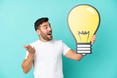 Mavi arka planda izole edilmiş yakışıklı beyaz bir adam elinde bir ampul simgesi tutuyor.