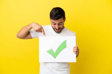 Sarı arka planda izole edilmiş yakışıklı beyaz bir adam elinde yeşil işaret simgesi olan bir pankart tutuyor ve onu işaret ediyor.