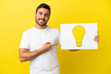 Sarı arka planda izole edilmiş yakışıklı beyaz bir adam elinde ampul simgesi olan bir pankart tutuyor ve onu işaret ediyor.