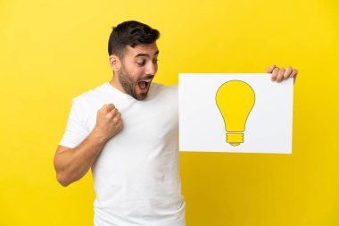 Sarı arka planda izole edilmiş yakışıklı beyaz adam elinde ampul simgesi olan bir levha tutuyor ve zaferi kutluyor.