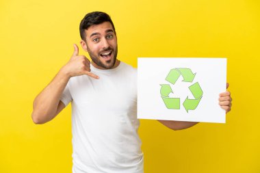 Sarı arka planda izole edilmiş yakışıklı beyaz adam elinde geri dönüşüm simgesi olan bir levha tutuyor ve telefon hareketi yapıyor.