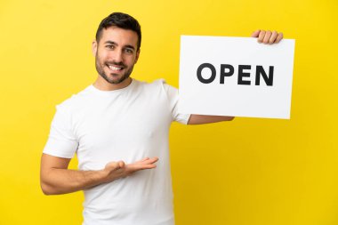 Sarı arka planda izole edilmiş yakışıklı beyaz bir adam. Elinde açık yazılı bir levha var ve onu gösteriyor.