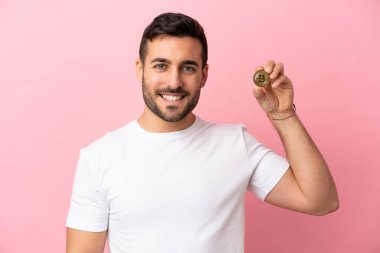 Elinde pembe arka planda izole edilmiş Bitcoin tutan genç adam.