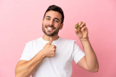 Elinde pembe arka planda izole edilmiş Bitcoin tutan genç adam iyi niyet gösterisi yapıyor.