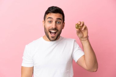 Elinde pembe arka planda, sürpriz bir yüz ifadesiyle izole edilmiş bir Bitcoin tutan genç adam.