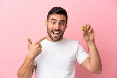 Elinde pembe arka planda izole edilmiş Bitcoin tutan genç adam iyi niyet gösterisi yapıyor.