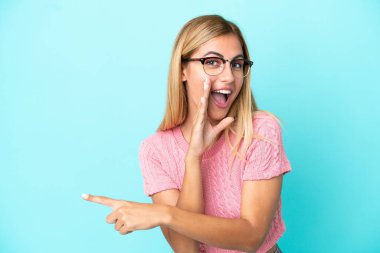Sarışın Uruguaylı kız mavi arka planda izole edilmiş bir şekilde bir ürün sunmak ve bir şeyler fısıldamak için kenarı işaret ediyor.